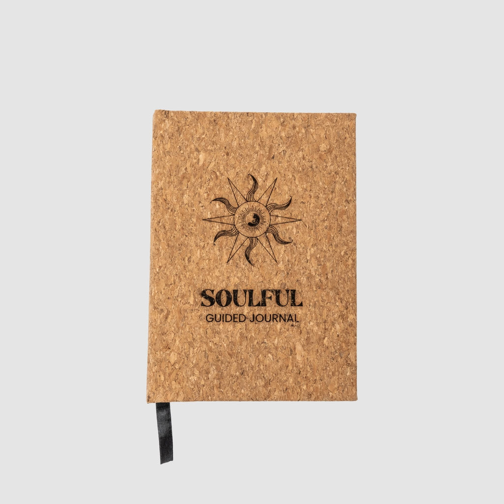 Cork-bound guided journal with round yin yang and ocean wave fused inspired logo, surrounded by a sun and star design, underneath titled 'Soulful Guided Journal' text all laser-engraved, sitting on a white background