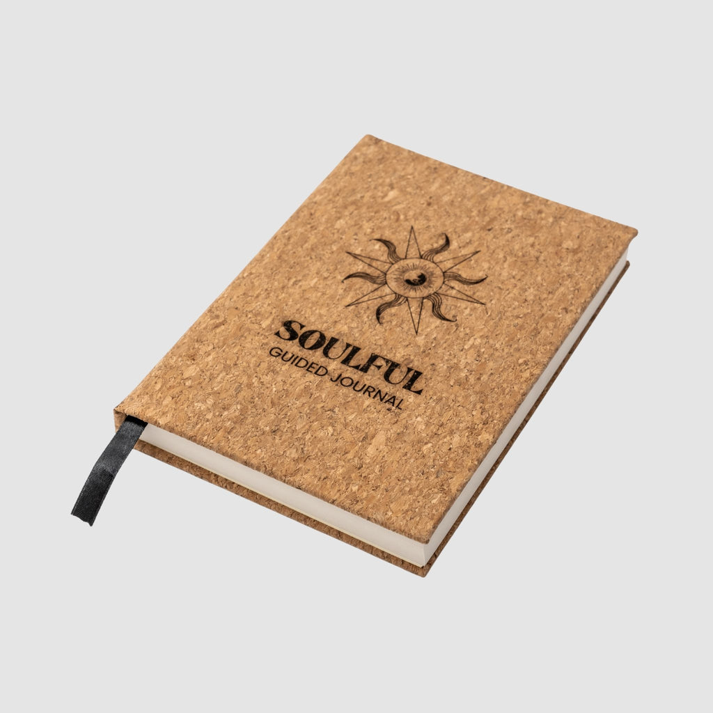 Cork-bound journal with 'Soulful Guided Journal' text and sun design sitting tilted on a white background