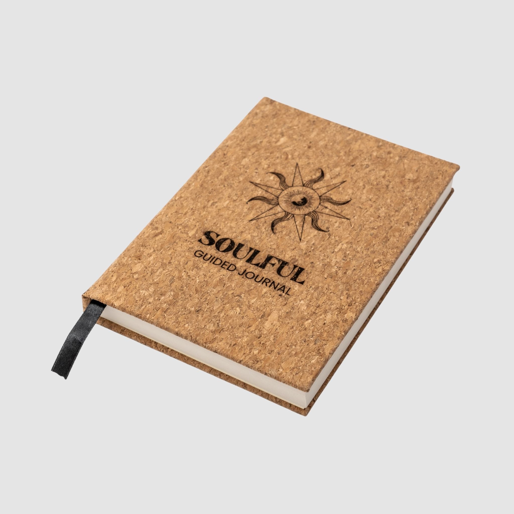 Cork-bound journal with 'Soulful Guided Journal' text and sun design sitting tilted on a white background