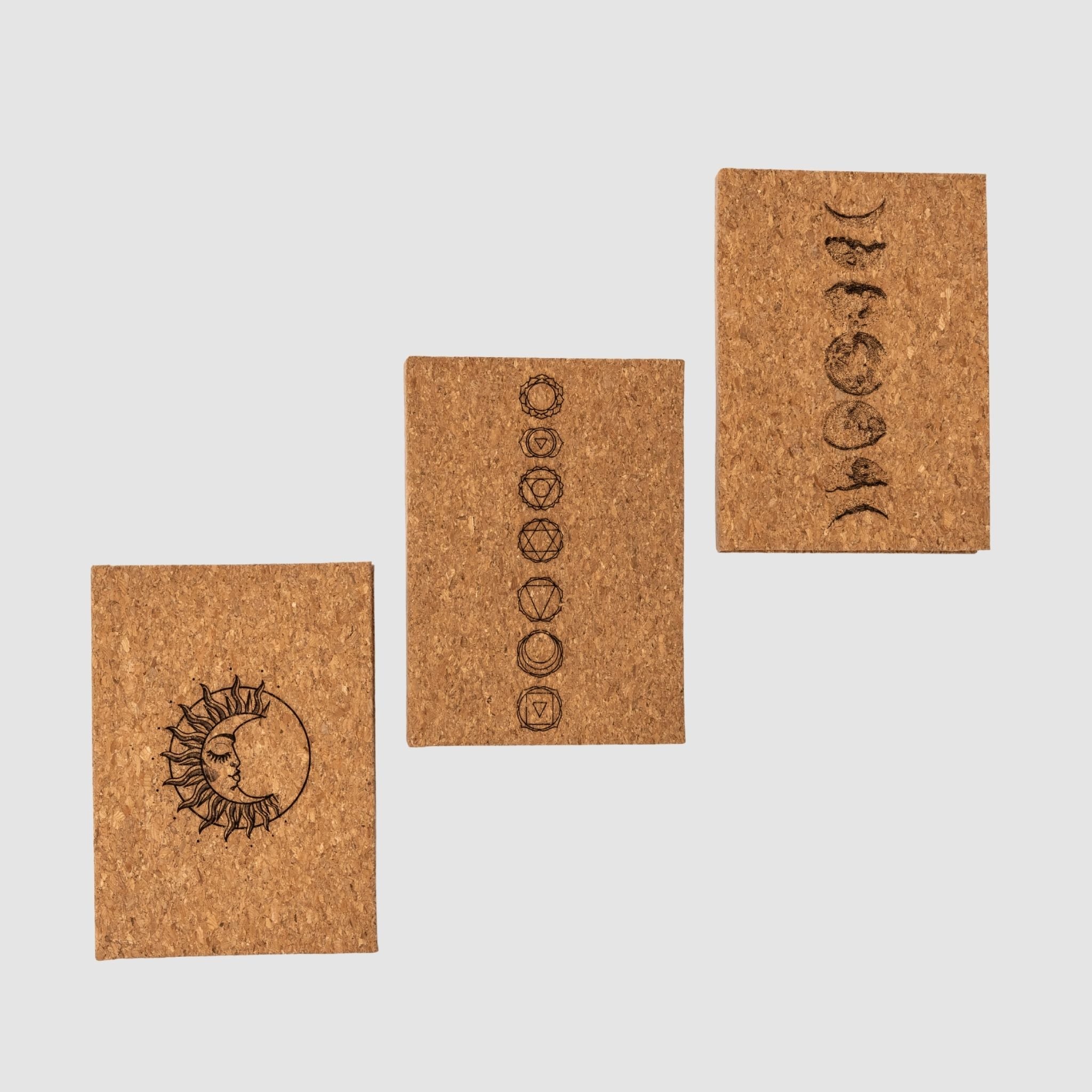 Three offset cork hardcover blank notebooks with sun-moon, chakra, and moon cycle designs, all on a white background