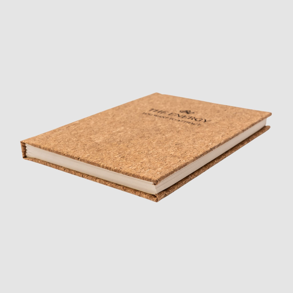 Cork-based planner with 'Be The Energy You Want To Attract' text laser-engraved design on front, sitting tilted on its side to show thickness, on a white background