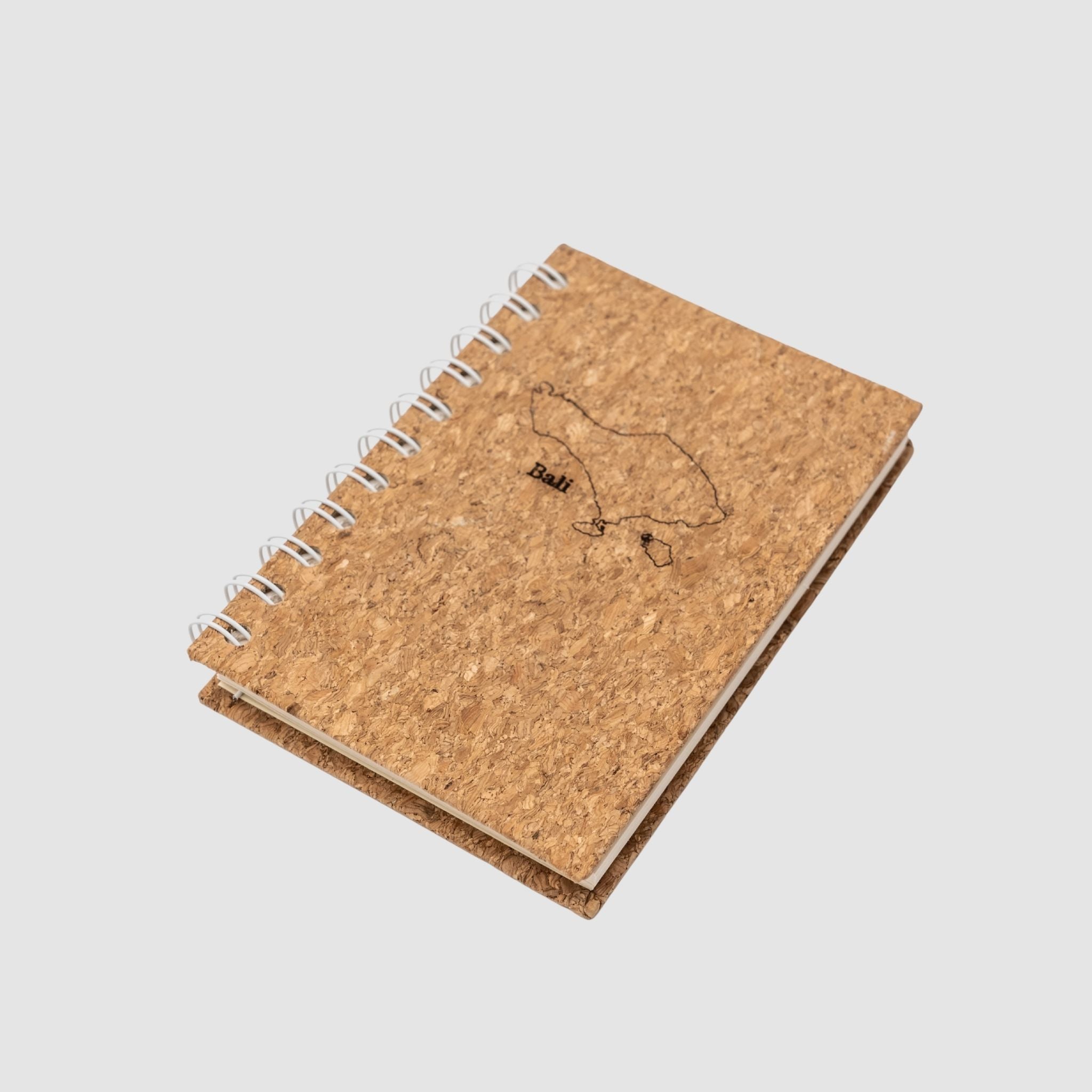 Small cork-based, compact spiral notebook, with a minimalistic bali island outline and the small word 'Bali' nicely fitted under it, all laser-engraved, sitting on a white background