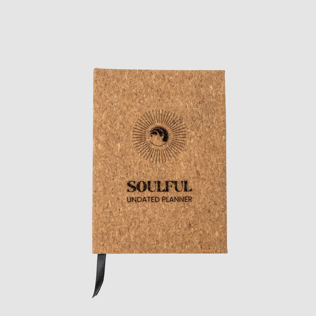 Cork planner with round yin yang and ocean wave fused inspired logo, surrounded by a circle sunburst, underneath titled 'Soulful Undated Planner' text all laser-engraved, sitting on a white background