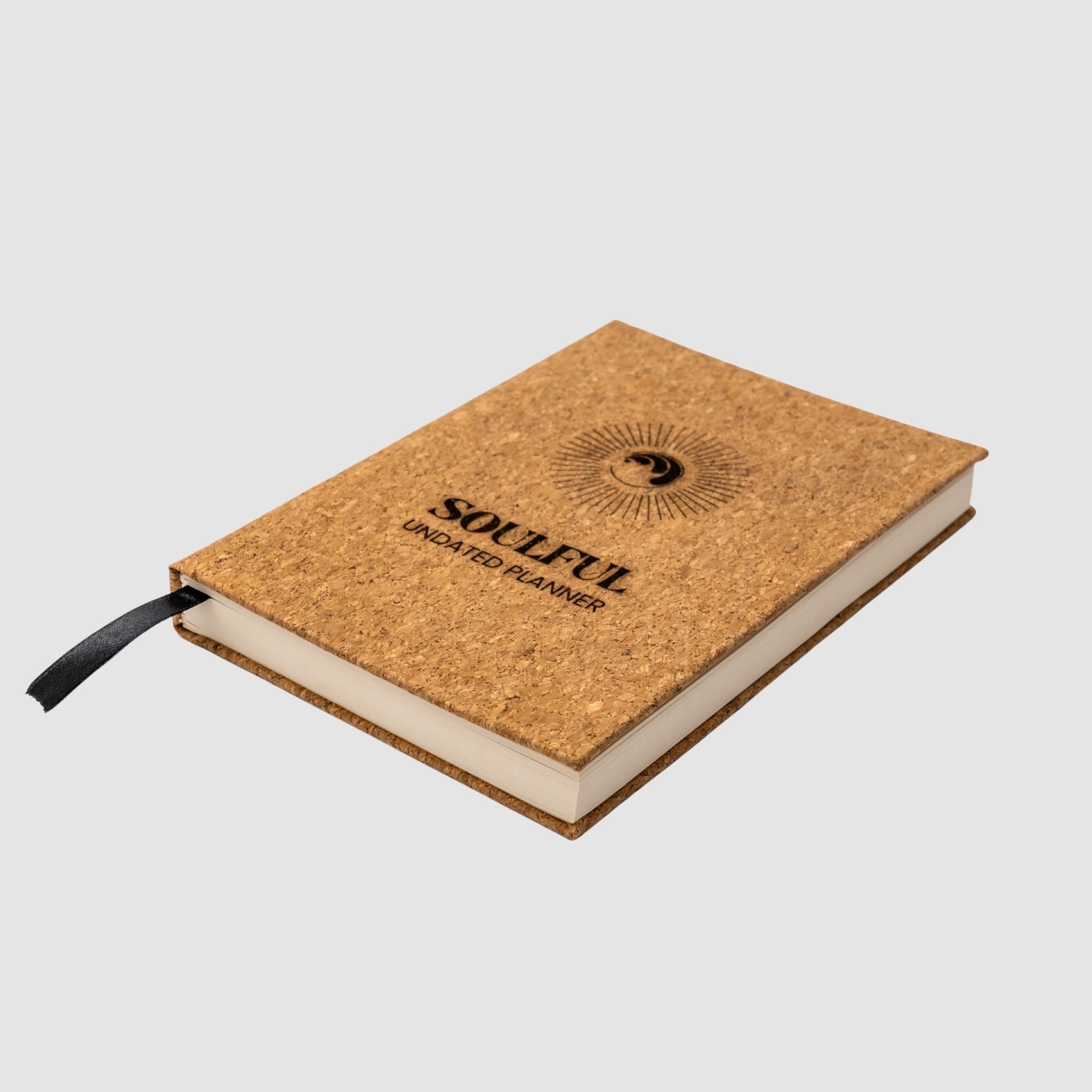 Cork planner with round yin yang and ocean wave fused inspired logo, surrounded by a circle sunburst, underneath titled 'Soulful Undated Planner' text all laser-engraved, sitting tilted a white background